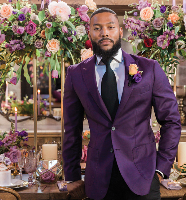 Trending groom suits groom wearing purple jacket posing for photo in front of large flower installations - groom trends