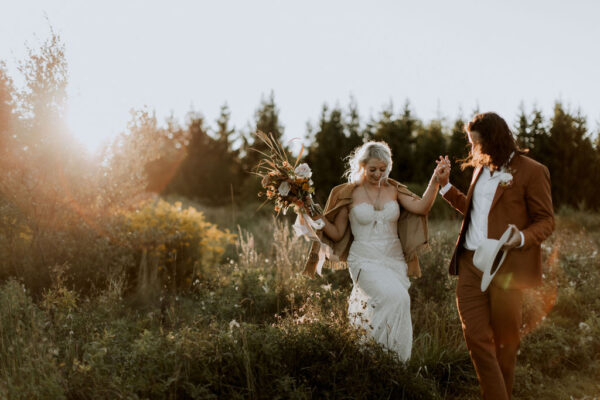 stylish groom wearing rust suit walking in field with his boho bride