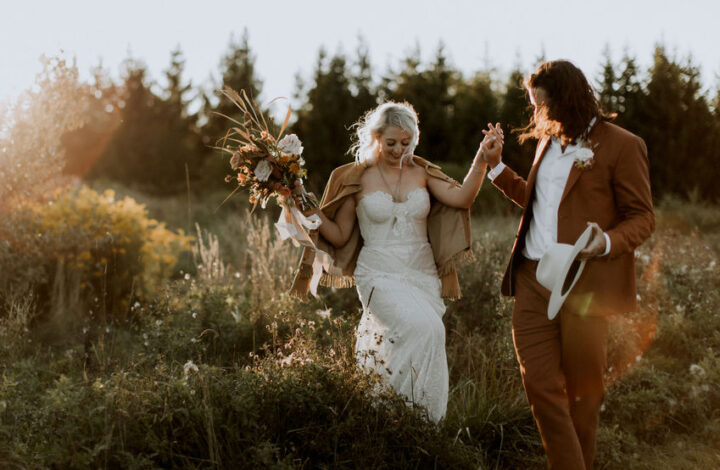 stylish groom wearing rust suit walking in field with his boho bride