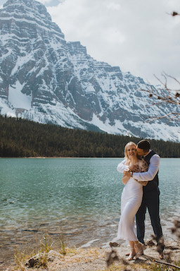 groom hugging and kissing bride next to Canadian lake with majestic mountain backdrop