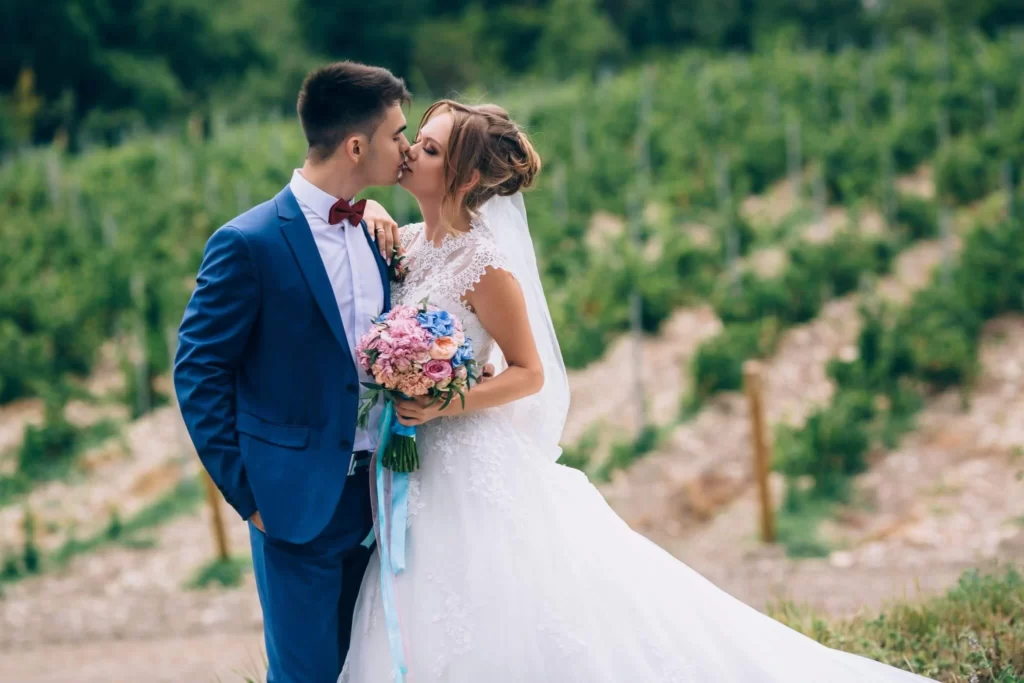groom wearing blue suit kissing bride in winery vineyard