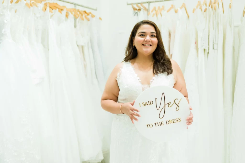 bride standing in front of wedding dress racks in bridal shop holding I said yes to the dress sign