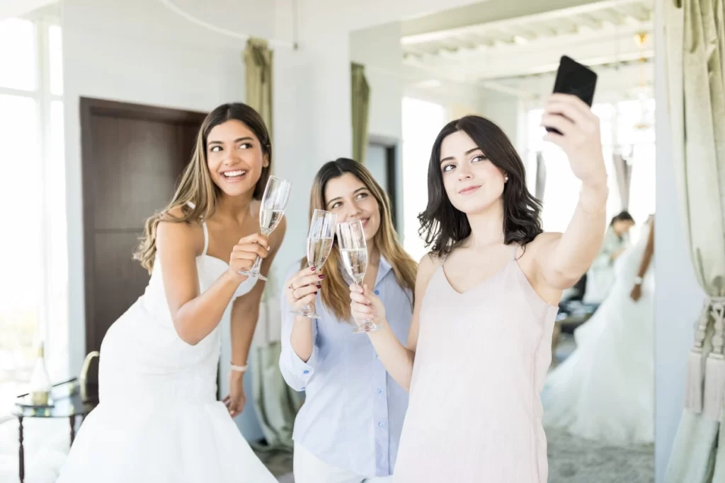 bridesmaid taking a selfie of bride and girls toasting champagne while trying on wedding dresses