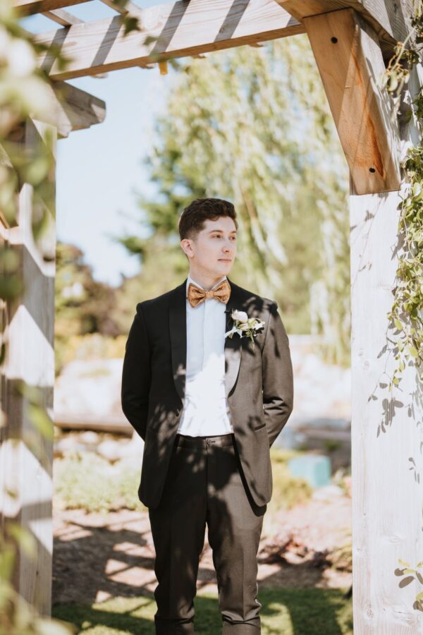 A groom, elegantly dressed in a tuxedo, stands gracefully beneath a beautifully adorned arbor.