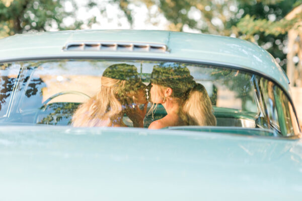 Two women sharing an affectionate kiss in the rear seat of a vintage automobile.