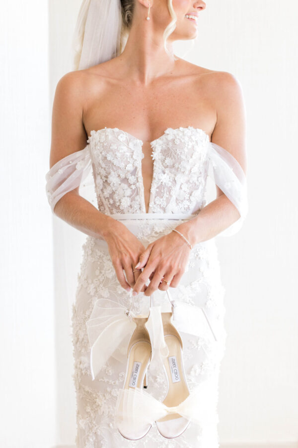 A beautiful bride in a white gown holds her wedding shoes, ready to walk down the aisle.