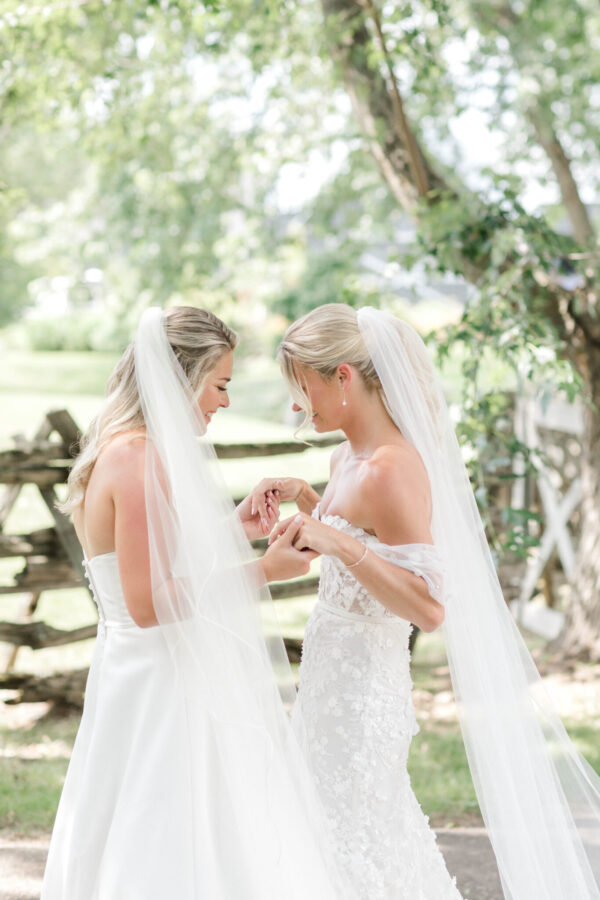 Two brides in elegant wedding dresses stand side by side, holding hands and smiling joyfully.