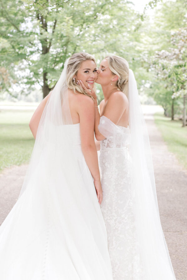 Two brides, dressed in elegant wedding gowns, share a tender kiss under the shade of a tree in a picturesque park.