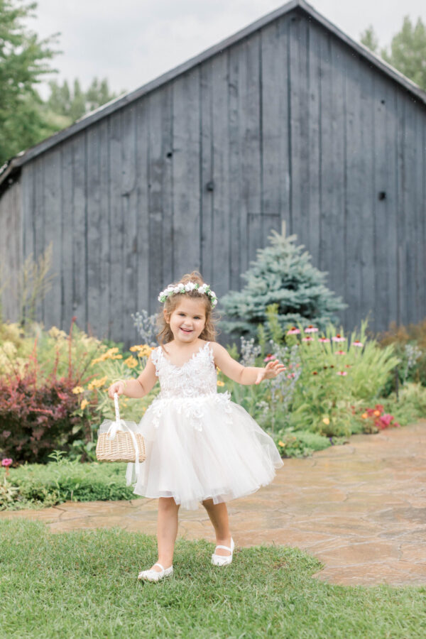A young girl wearing a white dress and tiara is walking in front of a rustic barn.