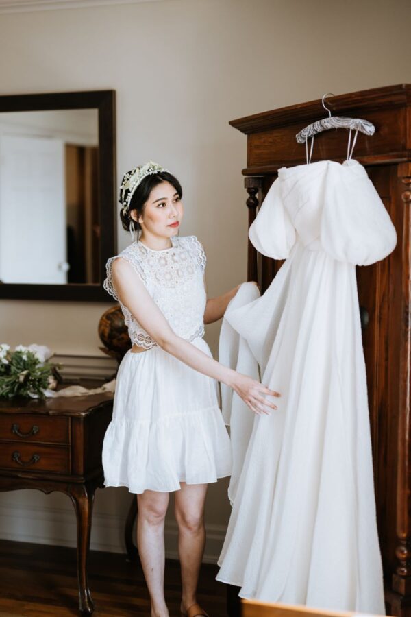A bride in a white dress gazes at her wedding gown