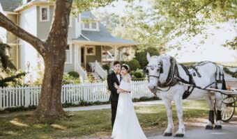 bride and groom pose in front of old house with horse drawn carriage