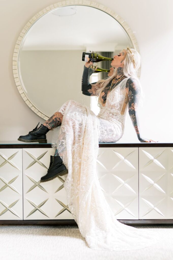  A bride elegantly seated on a table, adorned in a wedding gown, holding a bottle of wine.