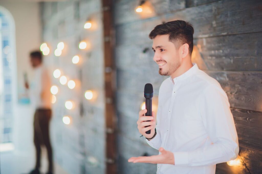 The best man is speaking into a microphone while dressed in formal attire, delivering a speech at a wedding reception.