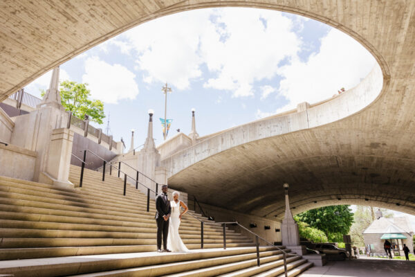 A couple, dressed in wedding attire, stands beneath a bridge, symbolizing their union and the start of their journey together.