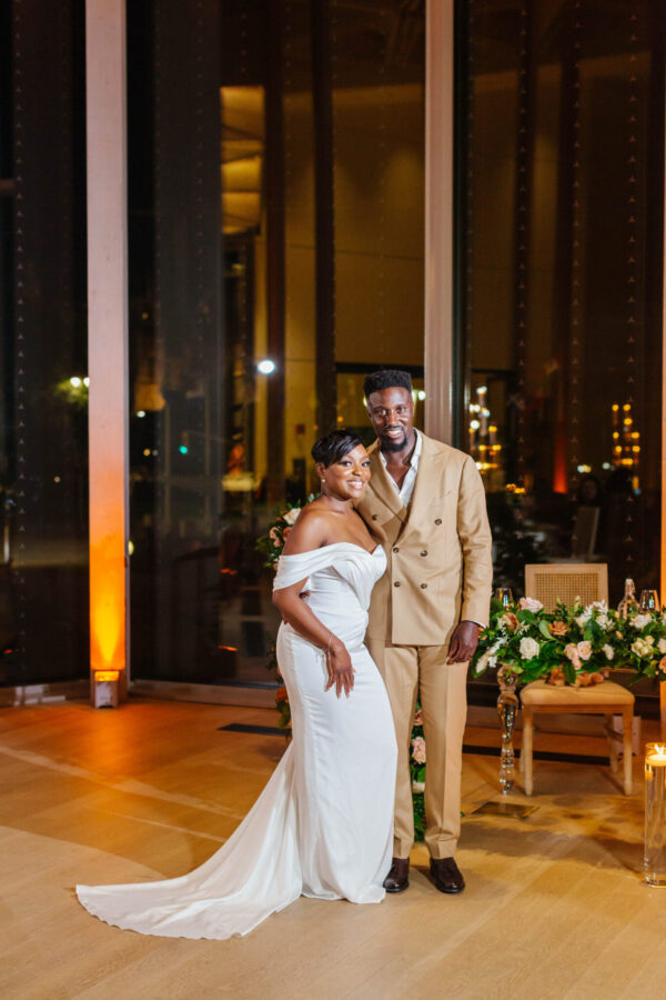 A bride and groom smiling, posing for a photo in front of a grand window, capturing their special day.