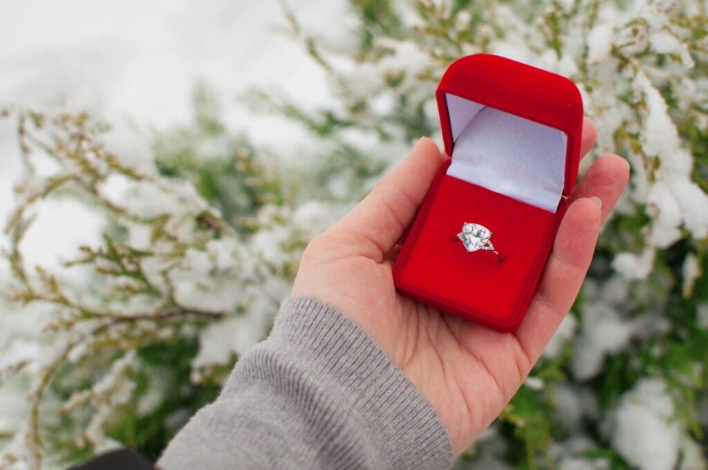 Chriistmas tree farm proposal engagement ring in red box with snowy Christmas tree in background winter wedding proposal