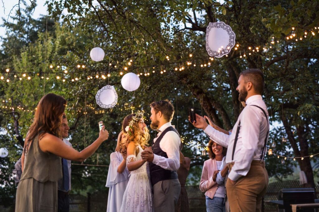 bride and groom dance under twinkle lights outside while wedding content creators take their photos on smartphones