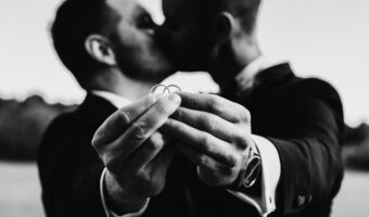black and white portrait of two grooms kissing and holding wedding rings together at destination wedding in France