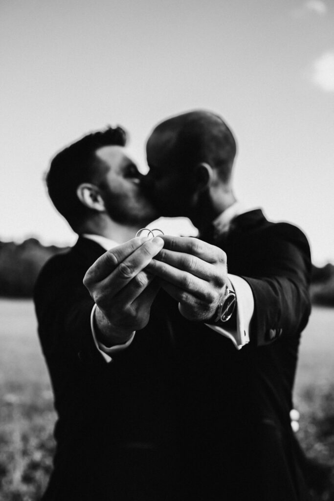 black and white portrait of two grooms kissing and holding wedding rings together at destination wedding in France