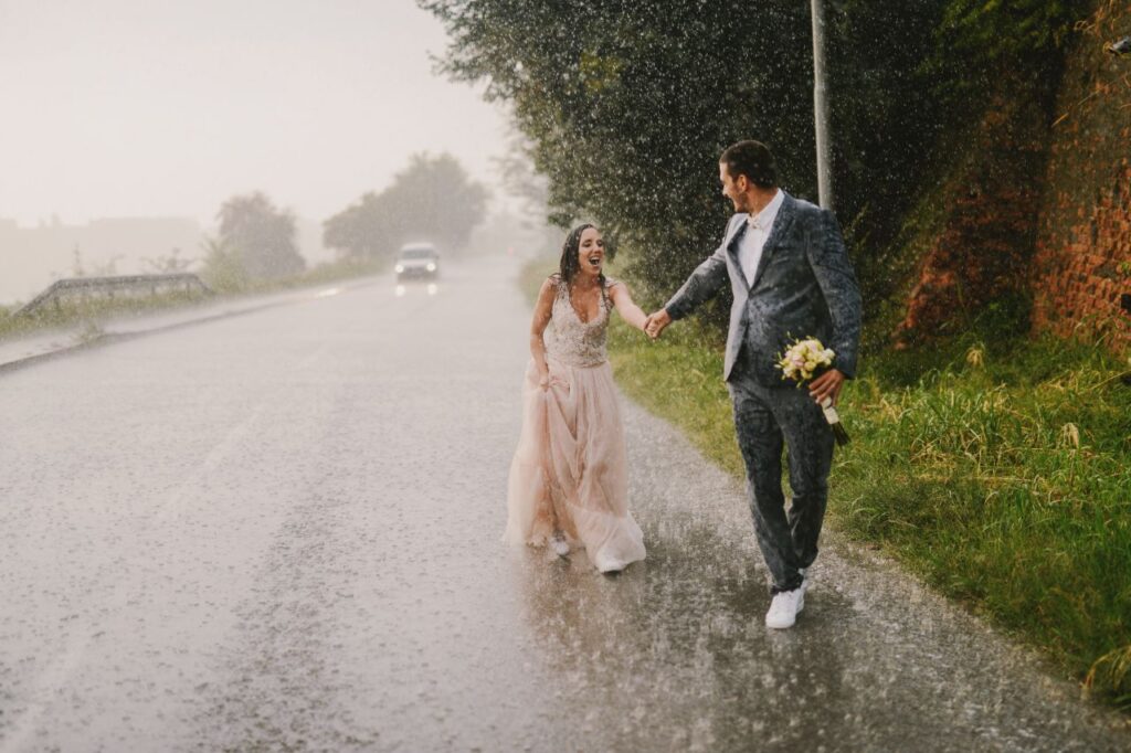 groom and bride hold hands while walking on road with rain on wedding day