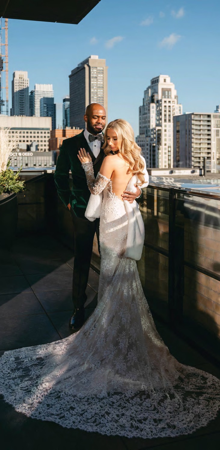 Bride in a lace wedding dress with a long train and a fur wrap embraces a man in a dark velvet suit on a rooftop with a city skyline behind them.