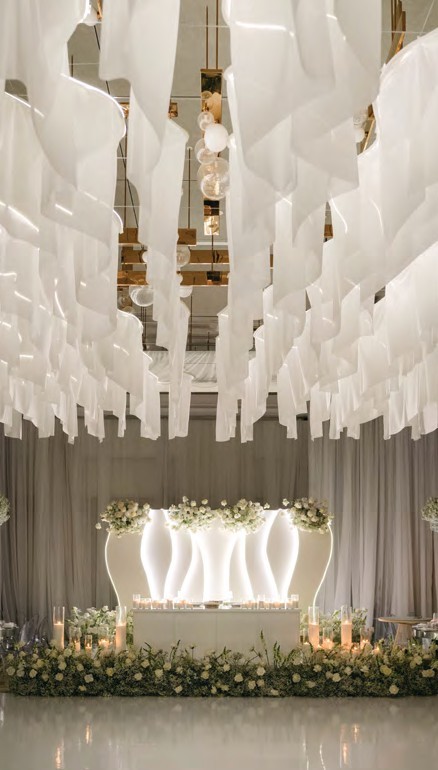 Wedding reception altar with flowing white fabric drapes, soft lighting, and a candlelit floral table above a greenery border.