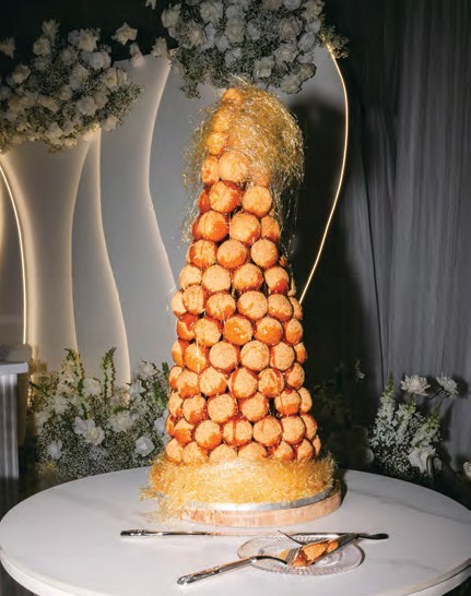 Tall pyramid of cream puffs stacked on a round pedestal, with gold sugar strands at the top, on a white-clothed table.