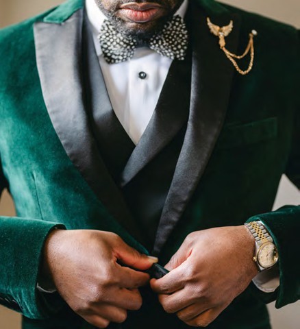 Man in a green velvet suit adjusts the jacket, wearing a patterned bow tie and a gold watch on his wrist.