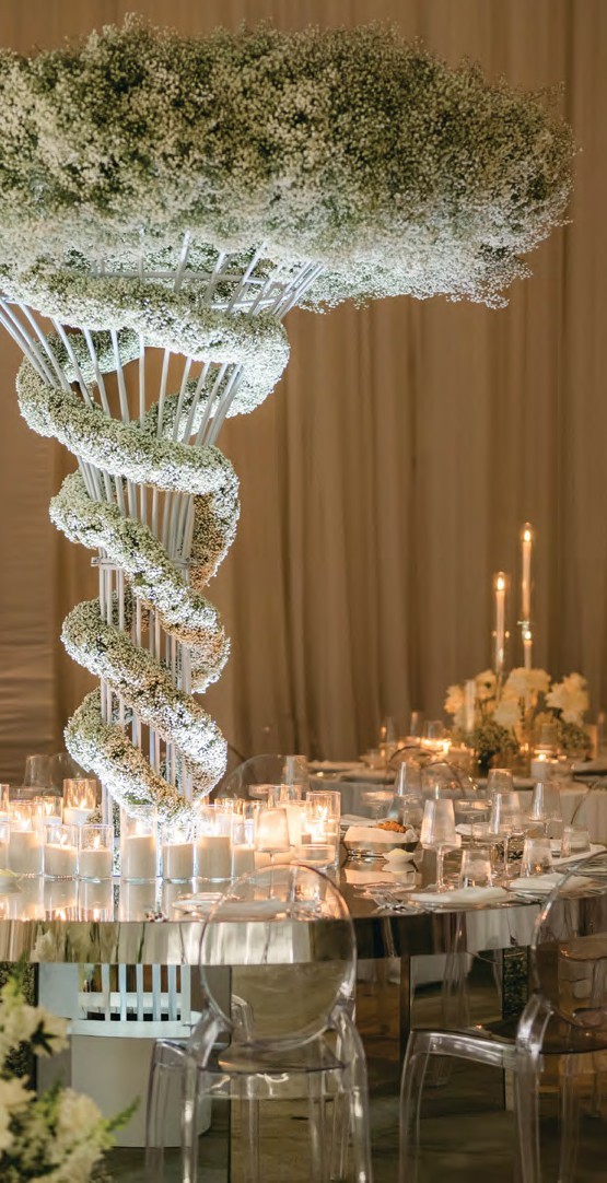 Elegant wedding reception table with a tall spiraling white floral centerpiece, candles, and clear chairs around a mirrored table surface.