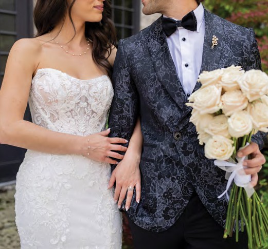 Bride in a white lace strapless gown and groom in a dark patterned suit arm in arm; the groom holds a bouquet of cream roses.