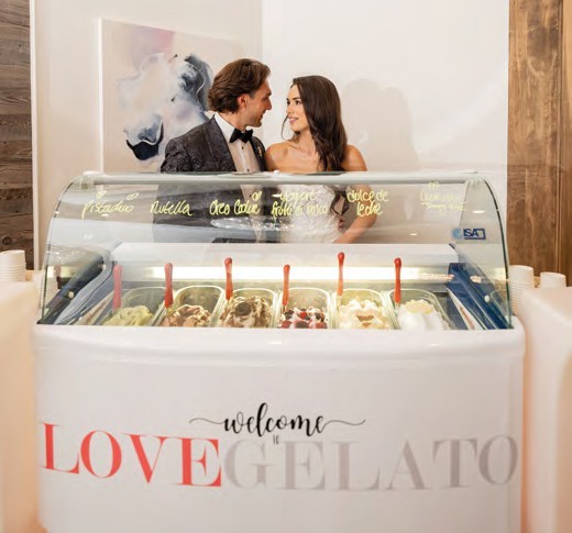 Bride and groom in wedding attire smile at each other as they stand behind a gelato counter with scoops on display and a front sign reading 'LOVE GELATO' .