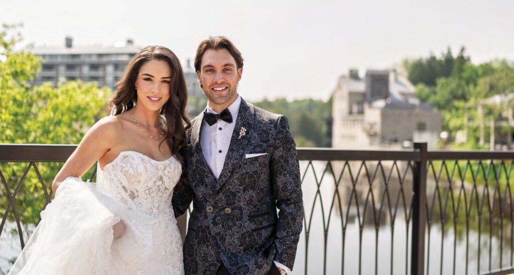Bride and groom posing together on a balcony by a river, smiling in wedding attire (dress and tuxedo).