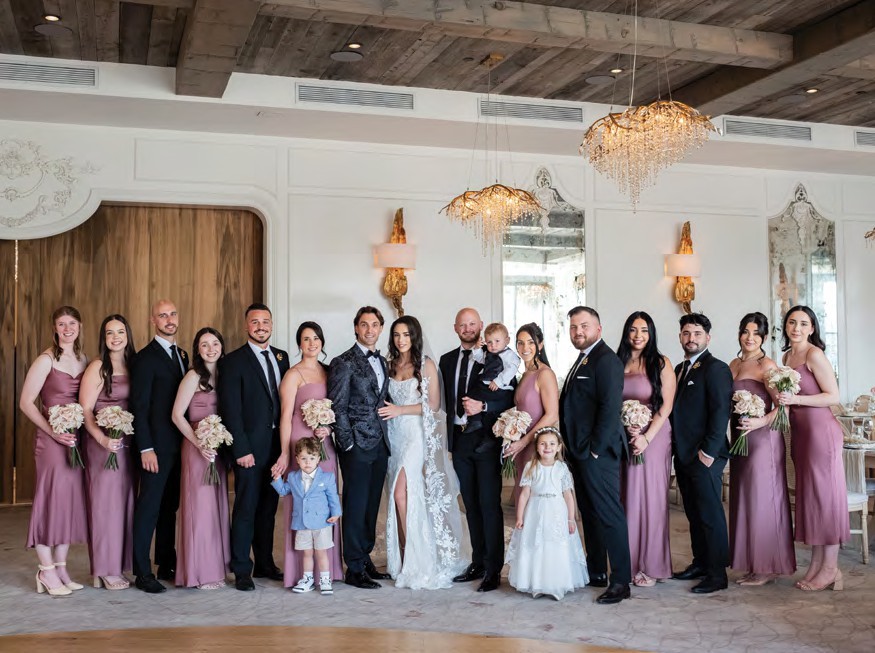 Wedding party photo: bride and groom center with bridesmaids in mauve dresses and groomsmen in suits, indoor venue with chandeliers.