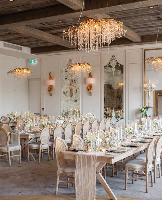 Elegant wedding reception hall with long wooden tables, crystal chandeliers, and gold-framed mirrors in a soft, cream palette.