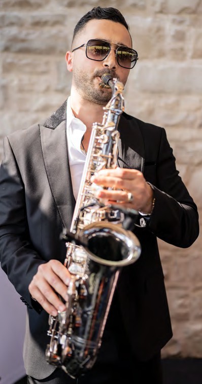 Man wearing a black suit and sunglasses playing a saxophone against a stone wall background.