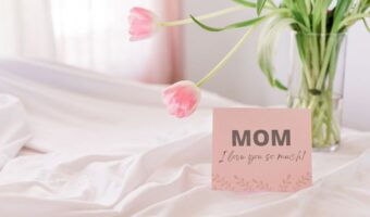 Pink greeting card on a white bed reads 'MOM I love you so much!' with a vase of pink tulips in the background.
