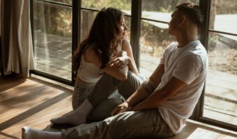 Couple sitting on a wooden floor by large windows, facing each other and chatting in a sunlit room