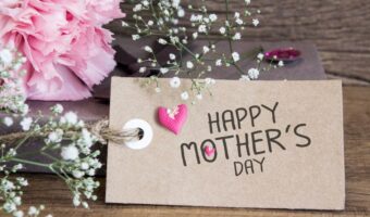 Brown craft tag reading 'Happy Mother's Day' with a pink heart, surrounded by pink carnations and white baby's breath on a wooden surface.