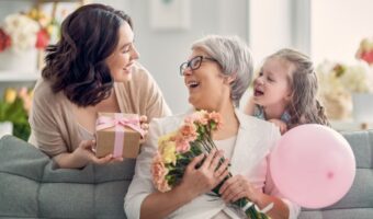 Three generations sharing a joyful moment on a sofa: an older woman with flowers, a younger woman presenting a gift, and a smiling child with a pink balloon behind them.