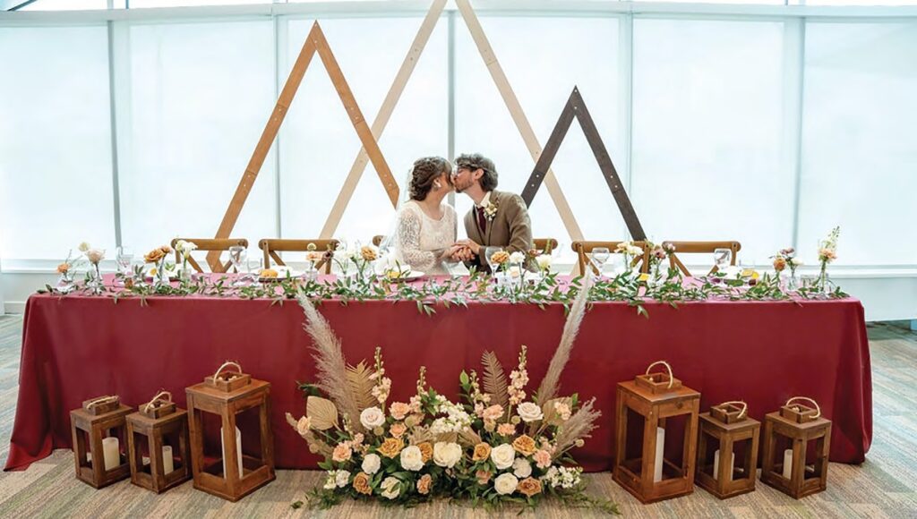 Bride and groom kiss at a long burgundy-draped head table adorned with pink and white flowers and greenery, with wooden lanterns on the floor nearby.