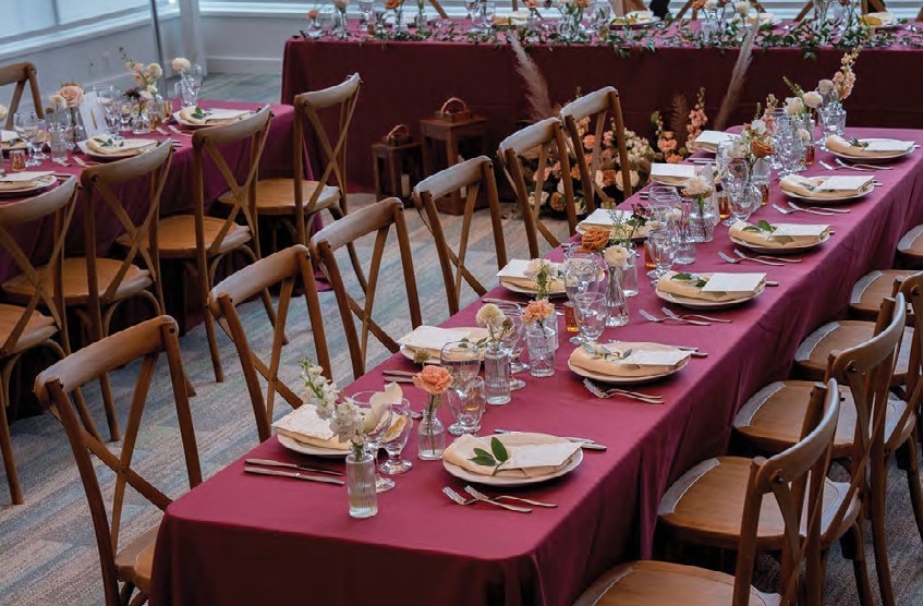 Long banquet table with burgundy tablecloths, place settings, and small floral centerpieces in a bright dining room.