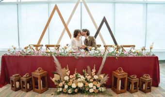 Bride and groom kiss at a long burgundy-draped head table adorned with pink and white flowers and greenery, with wooden lanterns on the floor nearby.