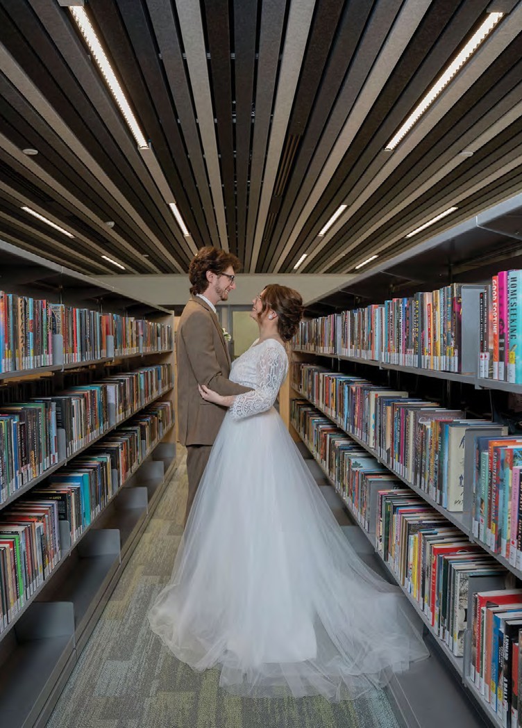 Bride in a white lace wedding gown and groom in a tan suit embrace in a library aisle lined with colorful books on both sides.