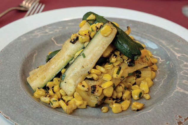 Grilled corn kernels and thick zucchini sticks, garnished with herbs, on a gray plate with a decorative rim.