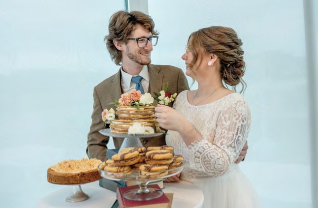 Bride and groom in wedding attire smile at each other while sharing a multi-tier dessert cake.