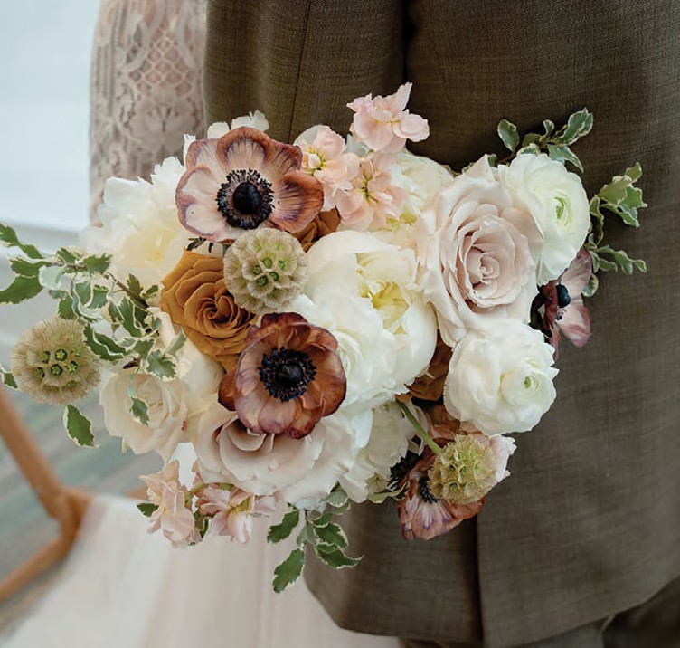 Bridal bouquet of white and blush roses with cream ranunculus and brown anemones, held by a person in a suit jacket