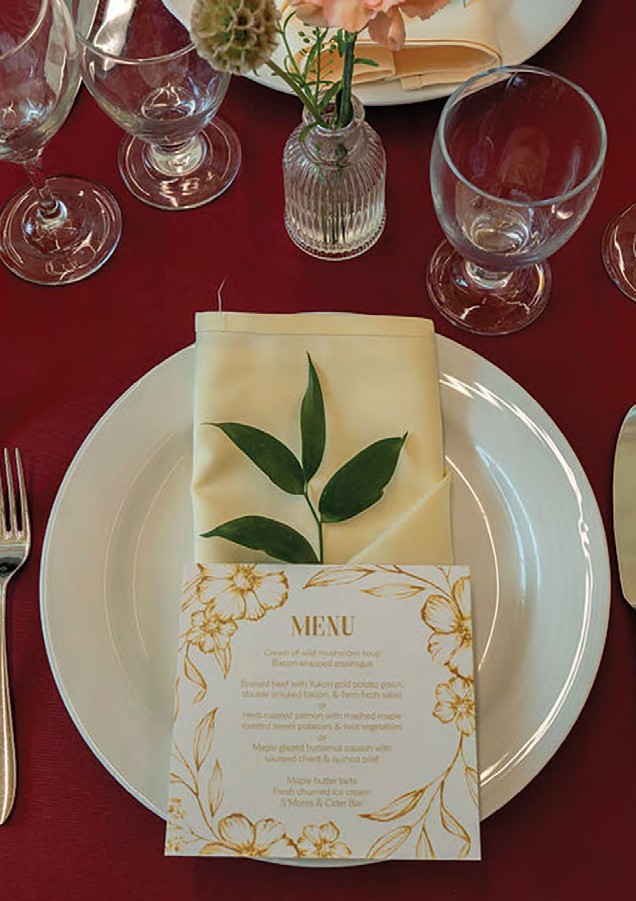 Table setting with a white plate, cream napkin folded with a green leaf, a floral menu card, and glassware on a burgundy tablecloth.