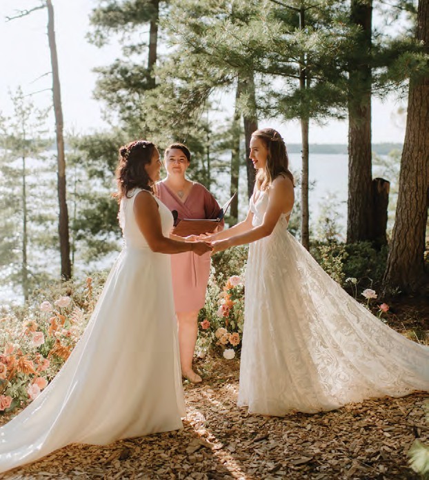 Two brides in white gowns exchange rings outdoors by a lakeside, with an officiant standing behind them among flowering bushes and pine trees.