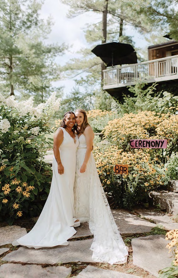 Two brides in white gowns pose on a stone path in a sunny garden with blooming flowers and a wooden deck nearby, signs read 'Ceremony' and 'Bar'.