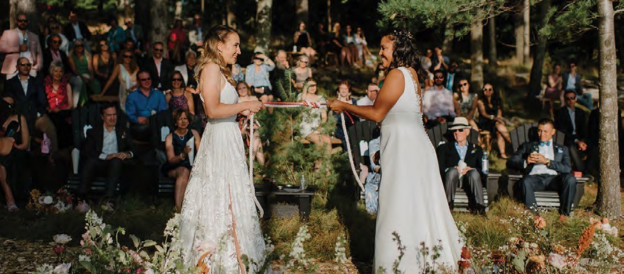 Two brides exchange ribbons in an outdoor wedding ceremony, surrounded by seated guests and trees.